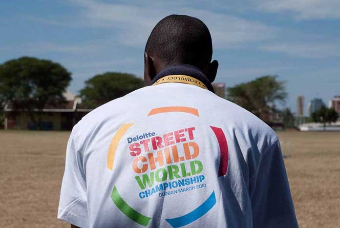 A man standing wearing a football shirt with the Street Child World Cup logo on the back.