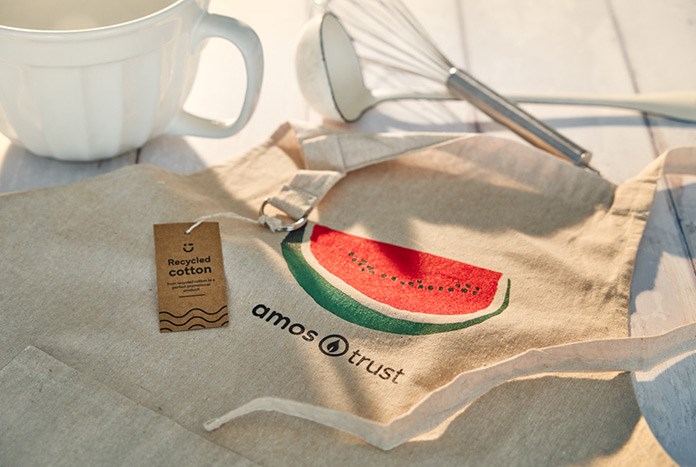An Amos Trust watermelon apron laying on a table with a bowl and egg wisk.