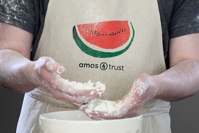 A man mixing flour in a bowl wearing an Amos Trust watermelon apron.