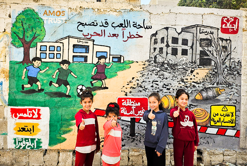 Four young children smiling in front of a mural in Gaza.