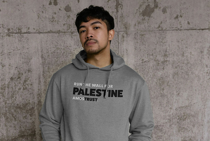 A young man wears a grey Amos Trust "I'm Running The Wall For Palestine' Hoodie.