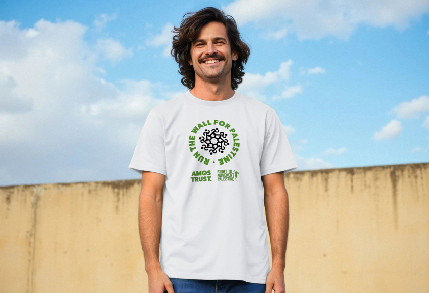 A man smiles for the camera wearing a white Amos Trust 'Run The Wall for Palestine T-shirt in front of a wall.